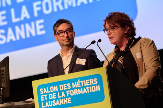 Un homme et une femme parlent sur un podium lors de l'événement du Salon des Métiers et de la Formation de Lausanne. Un grand panneau bleu est affiché à l'arrière-plan.