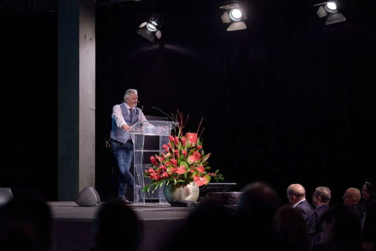 Un homme se tient debout sur un podium clair, sur une scène, et s'adresse à un public. Un grand bouquet de fleurs rouges et roses est posé à côté de l'estrade. Plusieurs hommes en costume sont assis au premier plan sous les lumières de la scène.