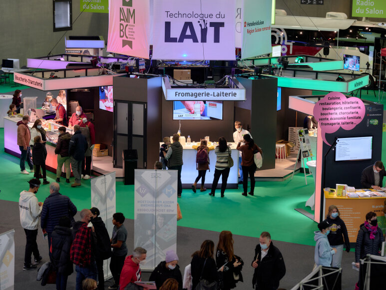 Un hall d'exposition très fréquenté présente des stands de technologie laitière et de fabrication de fromage. Des personnes se promènent, parcourent les stands et interagissent avec les exposants sous un éclairage coloré. Des panneaux et des bannières en français sont visibles tout au long de la scène.