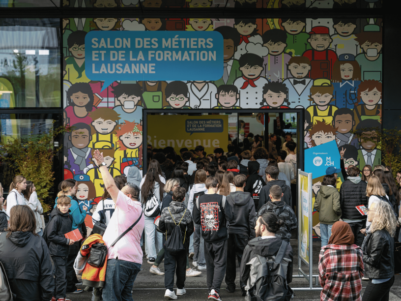 Une foule de personnes, principalement des étudiants, entre dans le bâtiment du Salon des Métiers et de la Formation de Lausanne. L'entrée est décorée de visages colorés de bandes dessinées et d'un grand panneau de bienvenue au-dessus des portes.