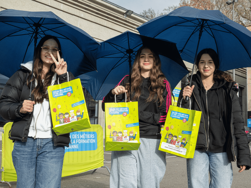 Trois jeunes femmes se tiennent à l'extérieur, tenant de grands parapluies bleus et des sacs cadeaux verts avec des dessins de bande dessinée, et souriant à la caméra. L'une d'elles fait un signe de paix avec sa main. Elles sont vêtues de vêtements décontractés et de vestes.