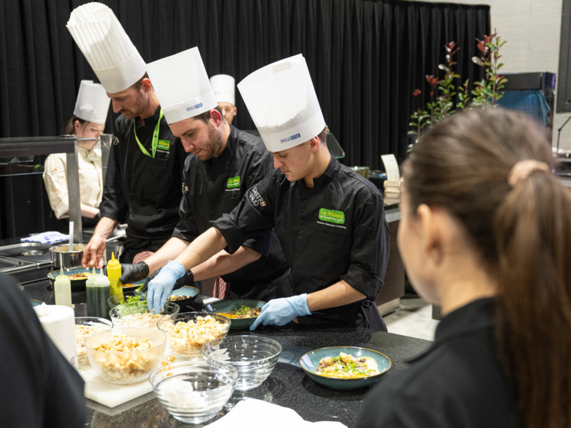Quatre chefs en uniformes noirs et chapeaux blancs préparent des plats au comptoir d'une cuisine, travaillant avec divers ingrédients et ustensiles, tandis qu'une femme au premier plan les observe.